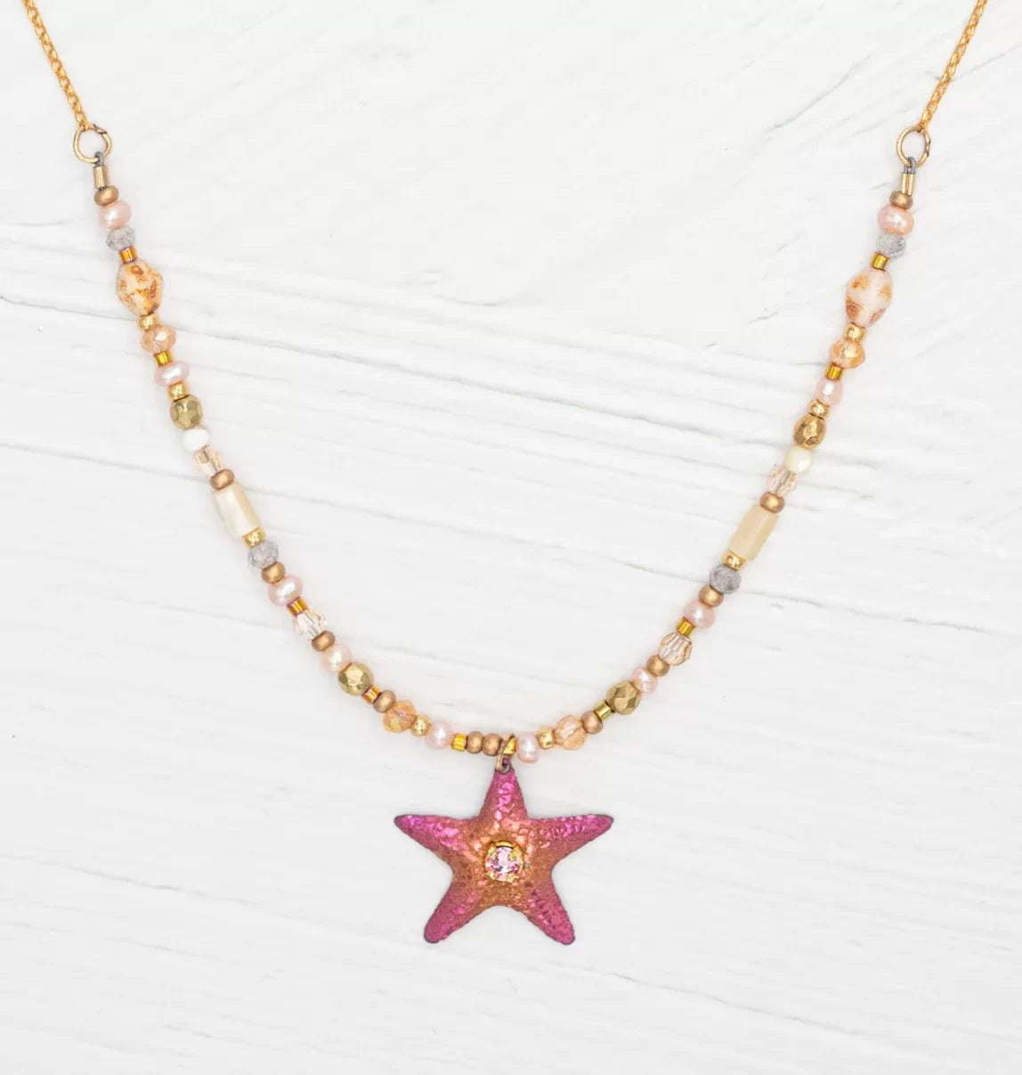 The Carmel Beaded Peach Starfish Necklace by Holly Yashi features a textured pink starfish pendant. Its chain, adorned with small, round beads in gold, pink, and beige shades, elegantly complements the serene beauty of the ocean against a light wooden background.