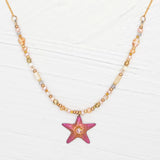 The Carmel Beaded Peach Starfish Necklace by Holly Yashi features a textured pink starfish pendant. Its chain, adorned with small, round beads in gold, pink, and beige shades, elegantly complements the serene beauty of the ocean against a light wooden background.