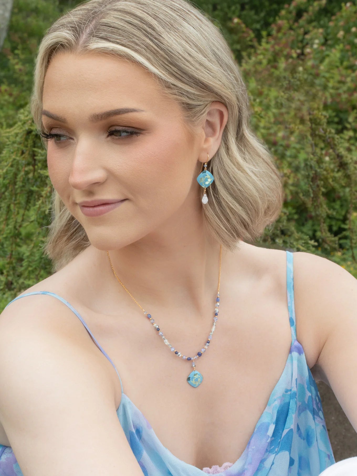A woman with short blonde hair wears the Elena Sky Blue Earrings by Holly Yashi, a blue patterned dress, and a gold necklace with a blue pendant. Outdoors among greenery, she gazes to the side with a gentle smile.