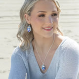 A woman with wavy blonde hair sits on a sandy beach, her smile radiant against the sea. She wears a light blue sweater, matching earrings, and her look is accentuated by the sparkling Austrian crystals of her Seashore Dreams Blue Depths Necklace from Holly Yashi Jewelry.