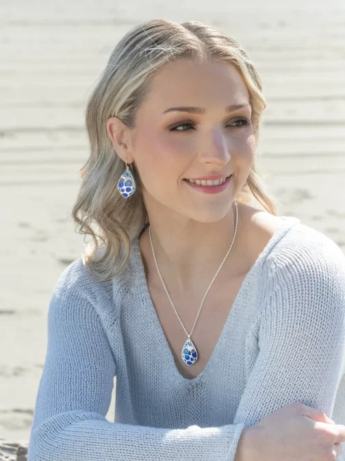 A woman with wavy blonde hair sits on a sandy beach, her smile radiant against the sea. She wears a light blue sweater, matching earrings, and her look is accentuated by the sparkling Austrian crystals of her Seashore Dreams Blue Depths Necklace from Holly Yashi Jewelry.