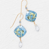 Elena Sky Blue Earrings by Holly Yashi feature gold hooks, blue floral square pendants, and sparkling crystal drops, displayed on a white textured background.