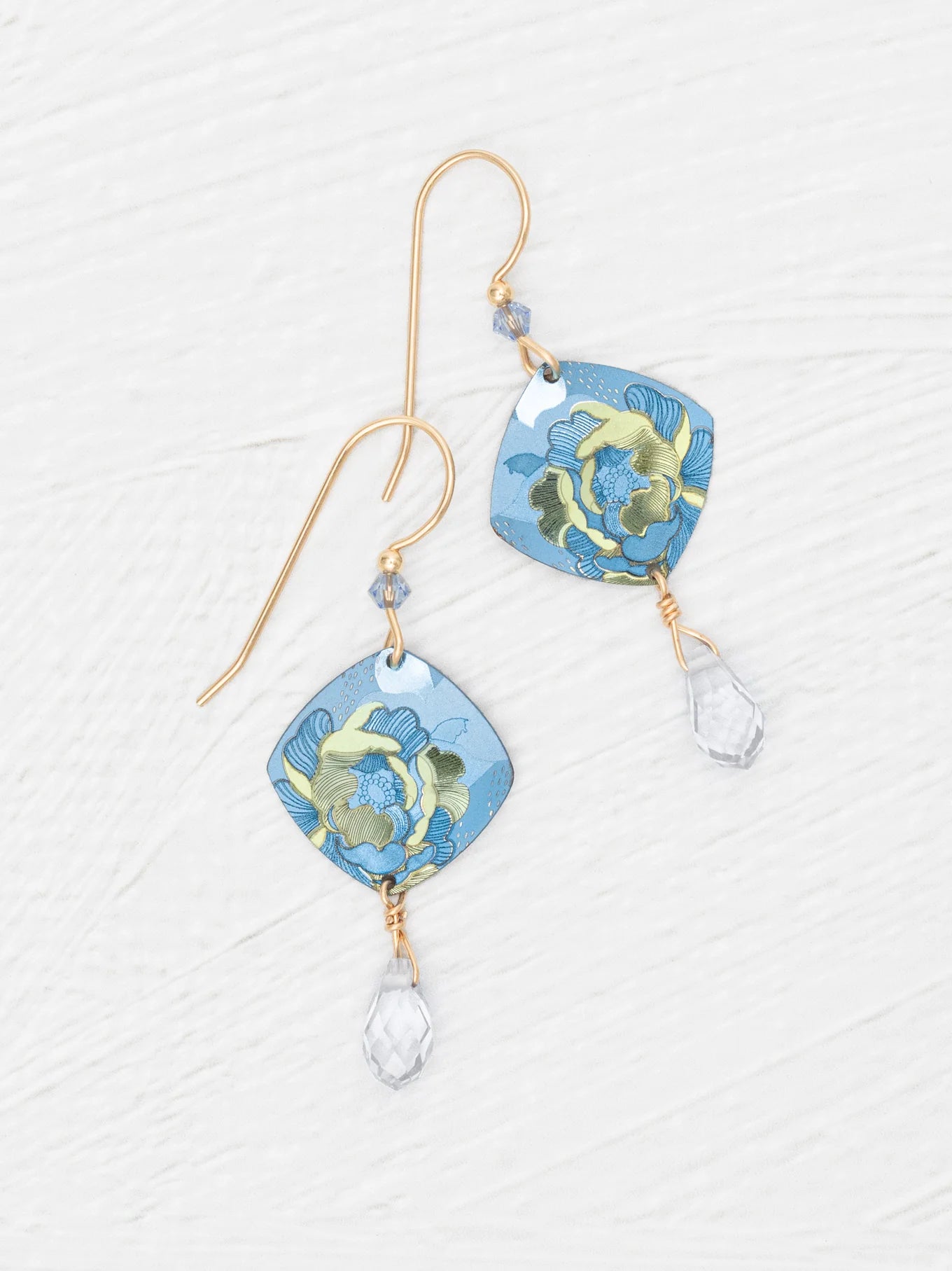 Elena Sky Blue Earrings by Holly Yashi feature gold hooks, blue floral square pendants, and sparkling crystal drops, displayed on a white textured background.