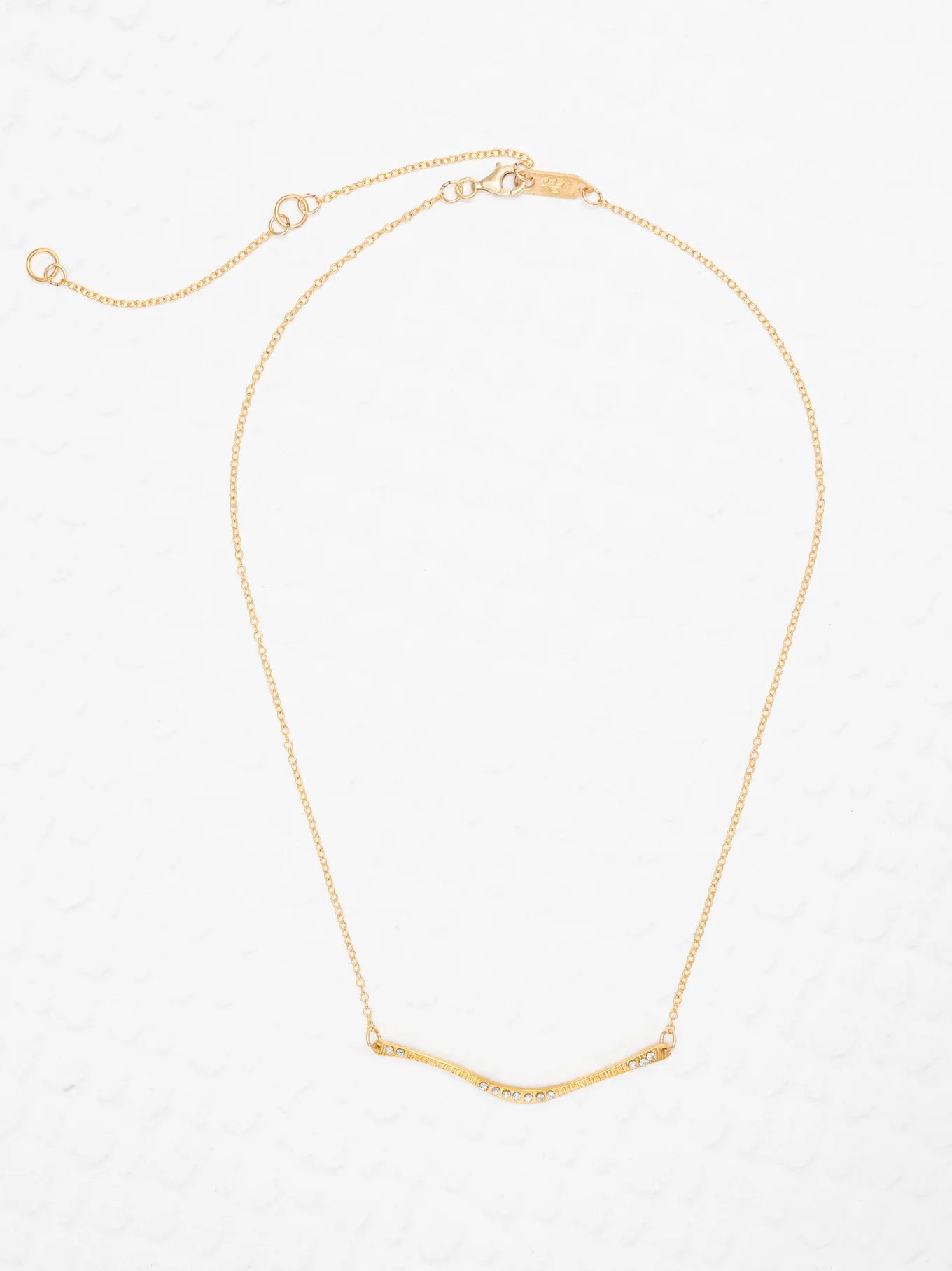 The Zara Bar Gold Necklace by Holly Yashi features a thin gold chain and curved bar pendant for a minimalist look. Hypoallergenic and stylish, this piece from Holly Yashi is shown on a white textured background.