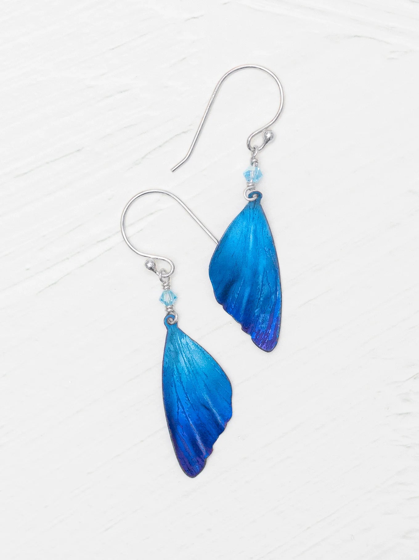 The Blue Flutterby Monarch Earrings by Holly Yashi are stunning, with translucent wing-shaped blue pendants featuring a deep to light blue gradient on silver hooks. Made of hypo-allergenic niobium, they beautifully mimic butterfly wings against a white textured surface.