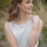 A woman in a white lace dress sits outdoors, smiling and looking to the side. She wears star-shaped earrings, a necklace, and her cherished Holly Yashi Carmel Beaded Seashore Blue Starfish Bracelet. The background is blurred with grass and greenery, echoing the peaceful essence of the oceans beauty.