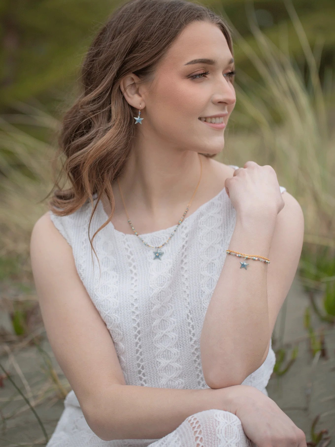 A woman in a white lace dress sits outdoors, smiling and looking to the side. She wears star-shaped earrings, a necklace, and her cherished Holly Yashi Carmel Beaded Seashore Blue Starfish Bracelet. The background is blurred with grass and greenery, echoing the peaceful essence of the oceans beauty.