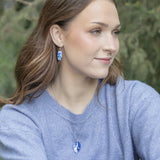 A woman with long brown hair, in a light blue sweater and matching necklace, wears Holly Yashi’s Falling Bloom Indigo Earrings, sitting outdoors against blurred greenery and gazing right with a gentle expression.