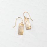 The Radiant Petra Gold Earrings by Holly Yashi are gold-plated with textured rectangular dangles and sparkling gemstone accents, elegantly displayed on a light patterned background.