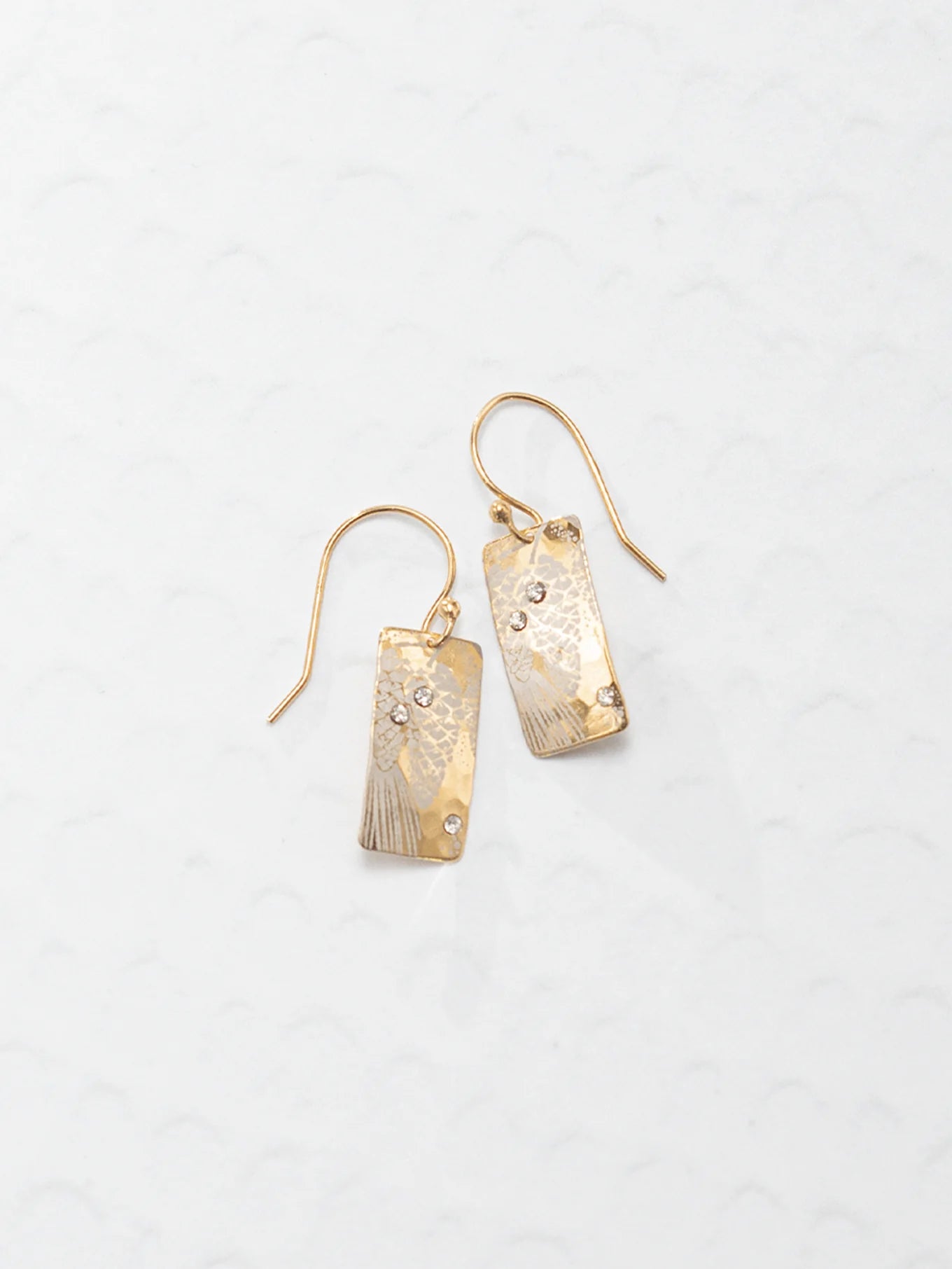 The Radiant Petra Gold Earrings by Holly Yashi are gold-plated with textured rectangular dangles and sparkling gemstone accents, elegantly displayed on a light patterned background.