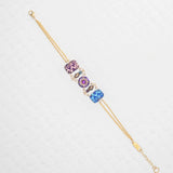The Alex Multi Color Bracelet by Holly Yashi features a gold chain with three jeweled art charms in purple, pink, and blue, each adorned with floral and swirl patterns, elegantly displayed on a textured white background.