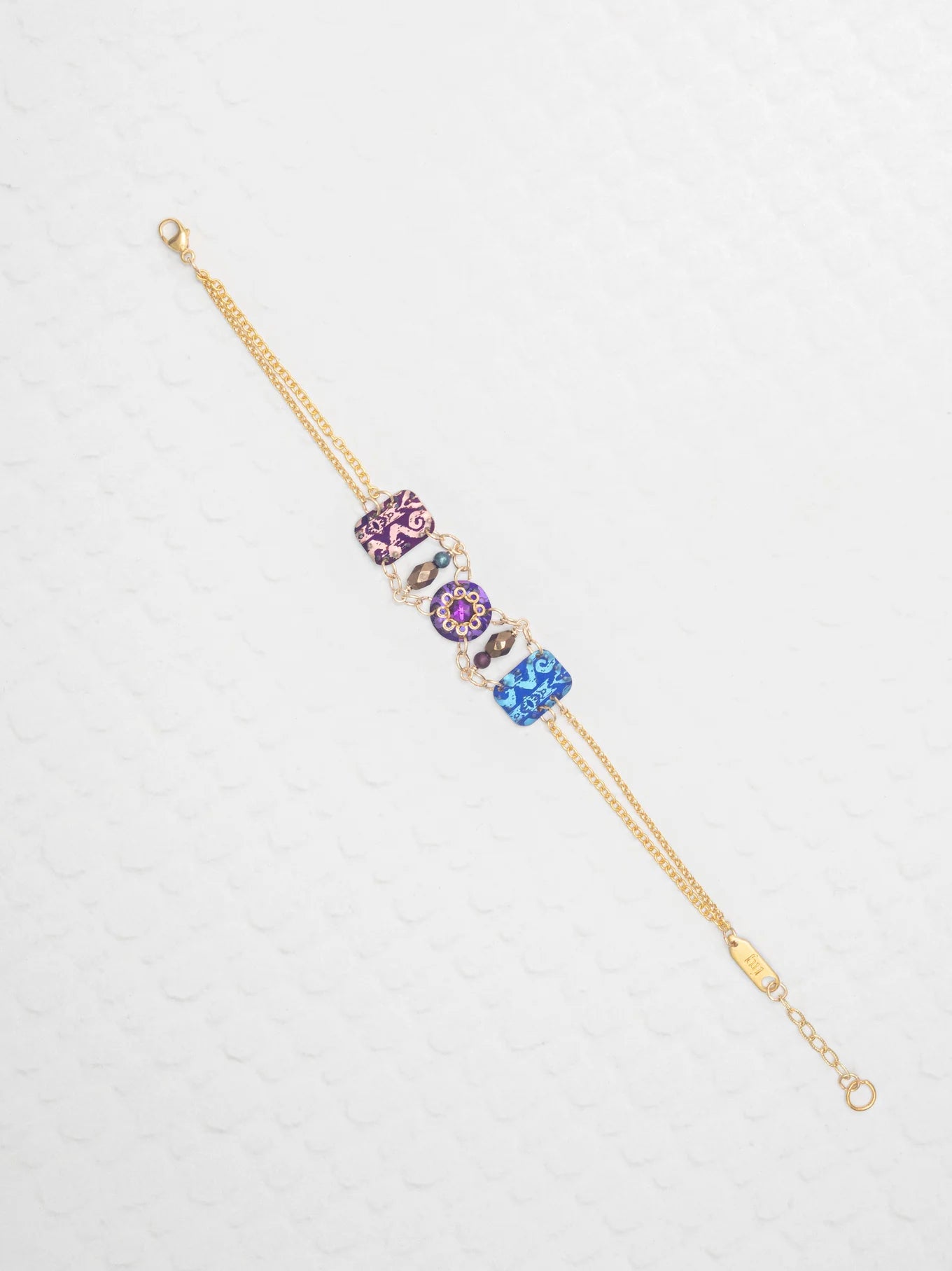 The Alex Multi Color Bracelet by Holly Yashi features a gold chain with three jeweled art charms in purple, pink, and blue, each adorned with floral and swirl patterns, elegantly displayed on a textured white background.