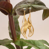 The Holly Yashi ER: Gilded Leaf Post Gold earrings feature sculptural leaf shapes with intricate cutouts, dangling elegantly from crystal posts and displayed on a large green plant leaf against a neutral background.