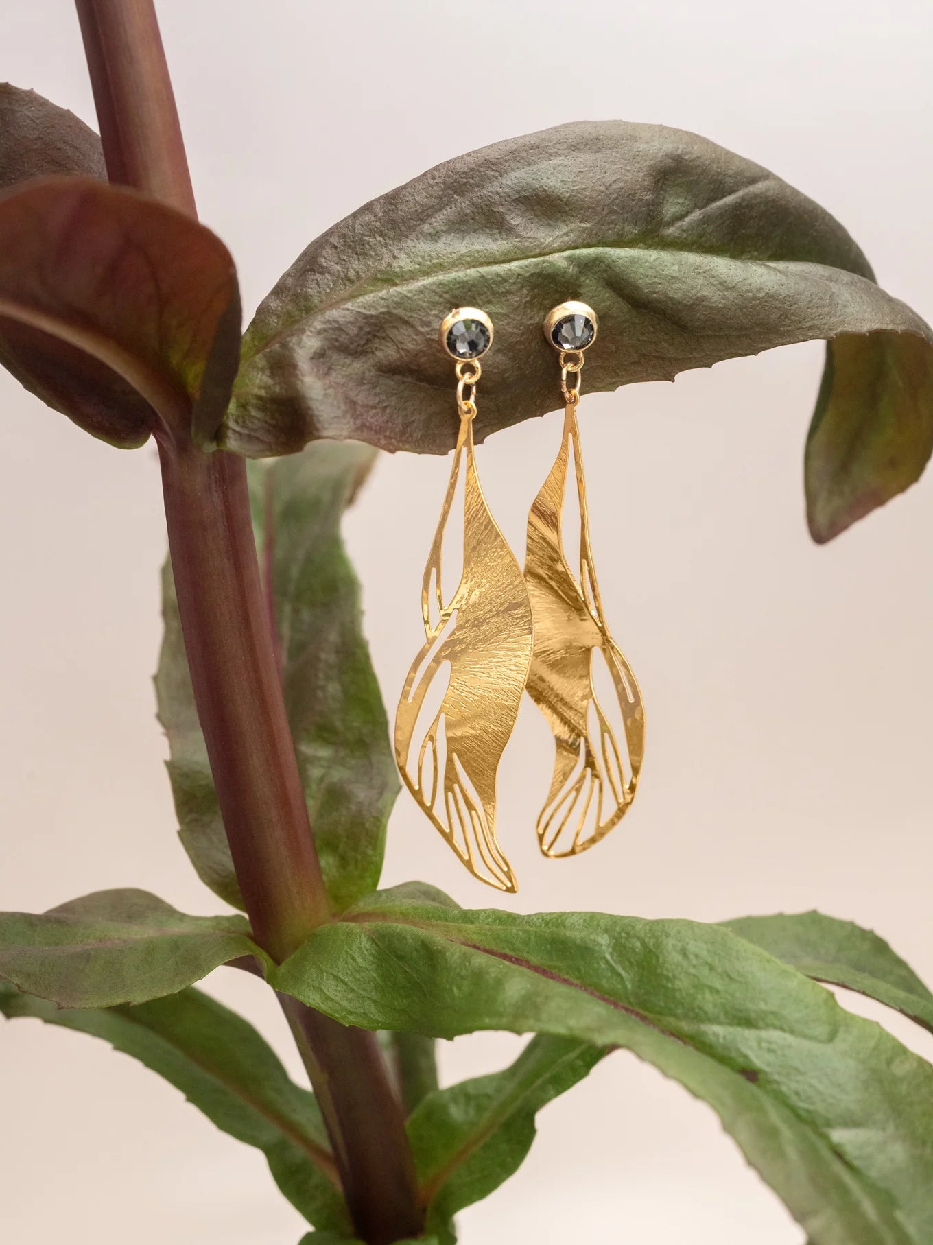 The Holly Yashi ER: Gilded Leaf Post Gold earrings feature sculptural leaf shapes with intricate cutouts, dangling elegantly from crystal posts and displayed on a large green plant leaf against a neutral background.