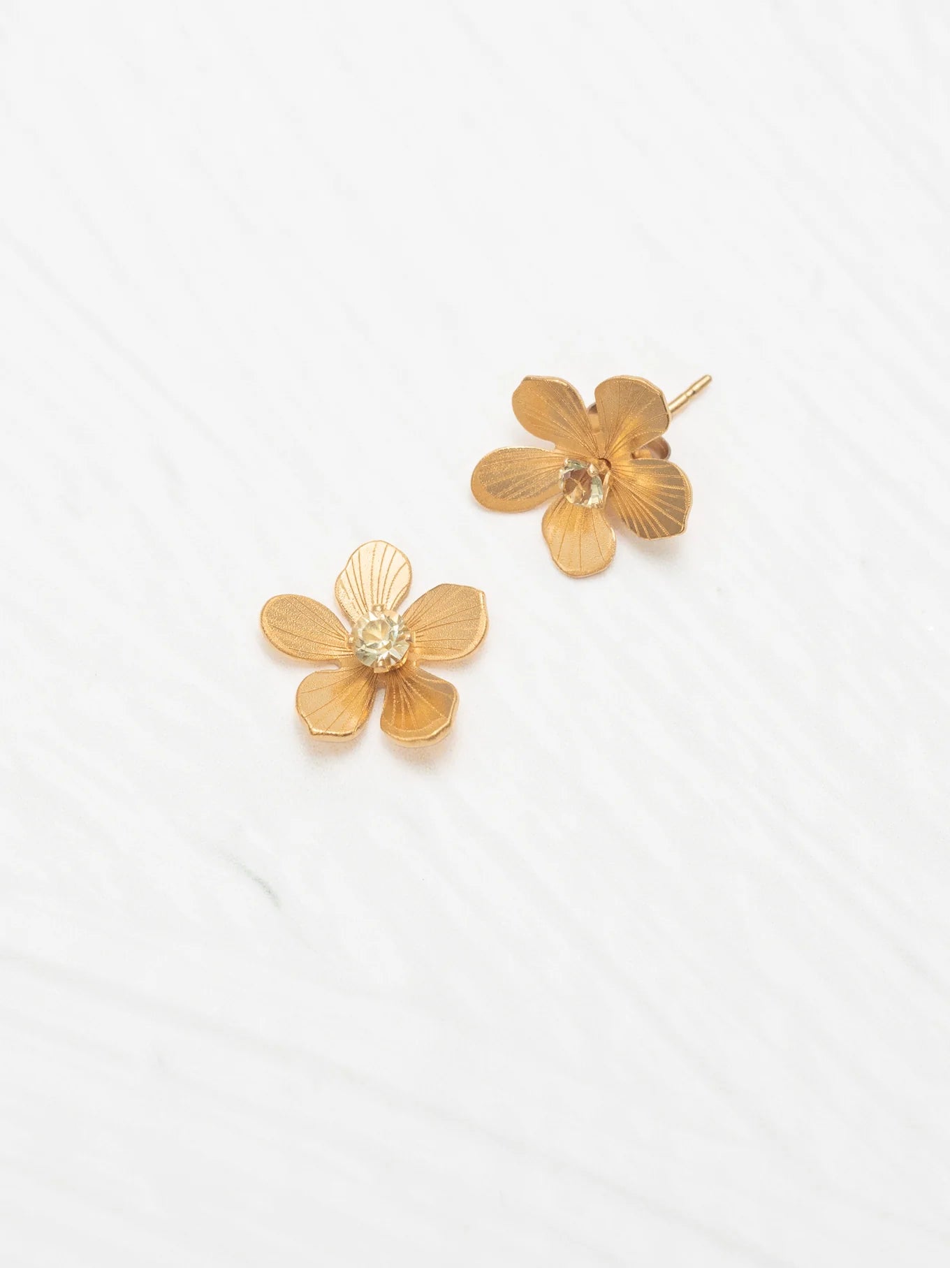 The Petite Plumeria Post Gold/Champagne Earrings by Holly Yashi feature textured flower-shaped studs with sparkling champagne centers, set on gold-filled posts—a charming accessory for any garden party, shown against a white textured background.