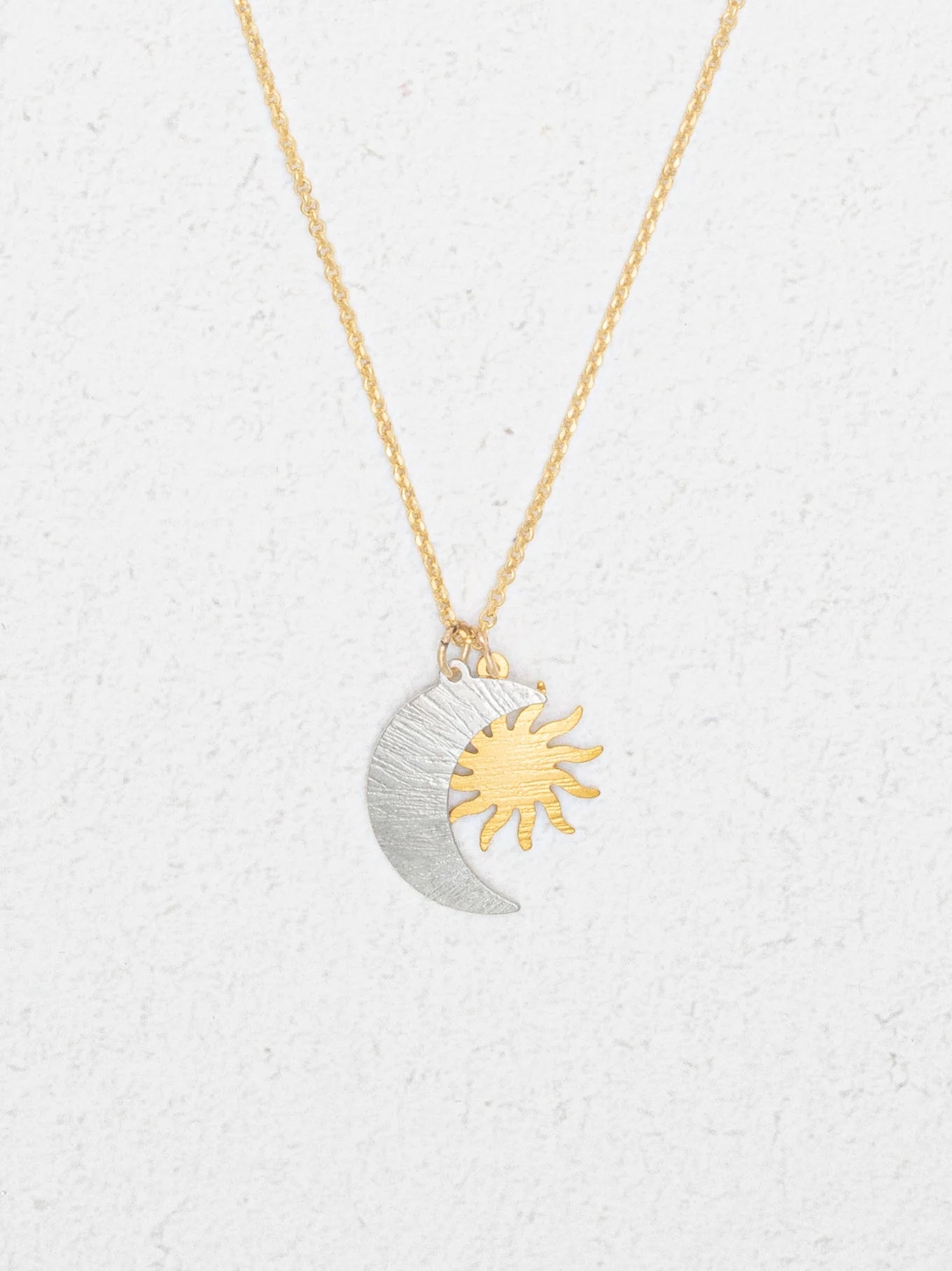 The Holly Yashi Sun and Moon Gold & Silver Necklace features overlapping silver crescent moon and gold sun pendants, reflecting a celestial jewelry style, displayed on a white textured background.