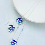 The Holly Yashi Bonnie Blue Garden Pansy Necklace, made from hypo-allergenic niobium, showcases silver pansy-shaped pendants in blue and white. Paired with matching earrings, this stunning set is elegantly displayed on a white circular platform against a light gray background.