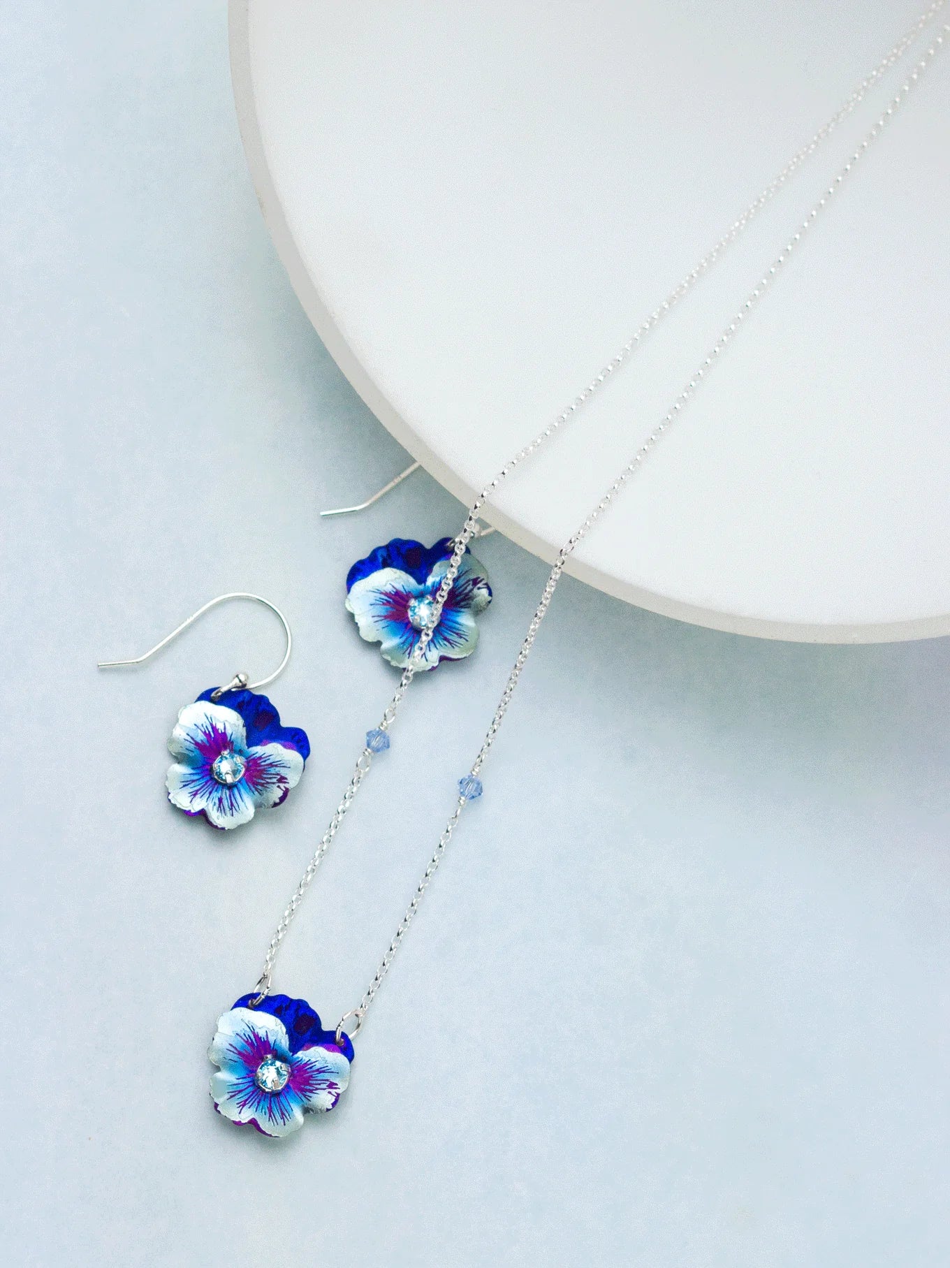 The Holly Yashi Bonnie Blue Garden Pansy Necklace, made from hypo-allergenic niobium, showcases silver pansy-shaped pendants in blue and white. Paired with matching earrings, this stunning set is elegantly displayed on a white circular platform against a light gray background.