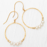 The Rosa Gold and Pearls Hoop Earrings by Holly Yashi feature a hammered texture with five freeform pearls at the bottom, hook fastenings, and are elegantly displayed on a white surface.