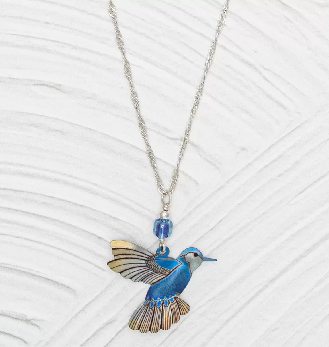 The Blue Picaflor Necklace by Holly Yashi features a vibrant blue and yellow hummingbird pendant crafted from hand-painted niobium, accented with a blue bead, all on an adjustable sterling silver chain against a textured light gray background.