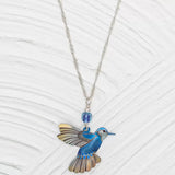 The Blue Picaflor Necklace by Holly Yashi features a vibrant blue and yellow hummingbird pendant crafted from hand-painted niobium, accented with a blue bead, all on an adjustable sterling silver chain against a textured light gray background.