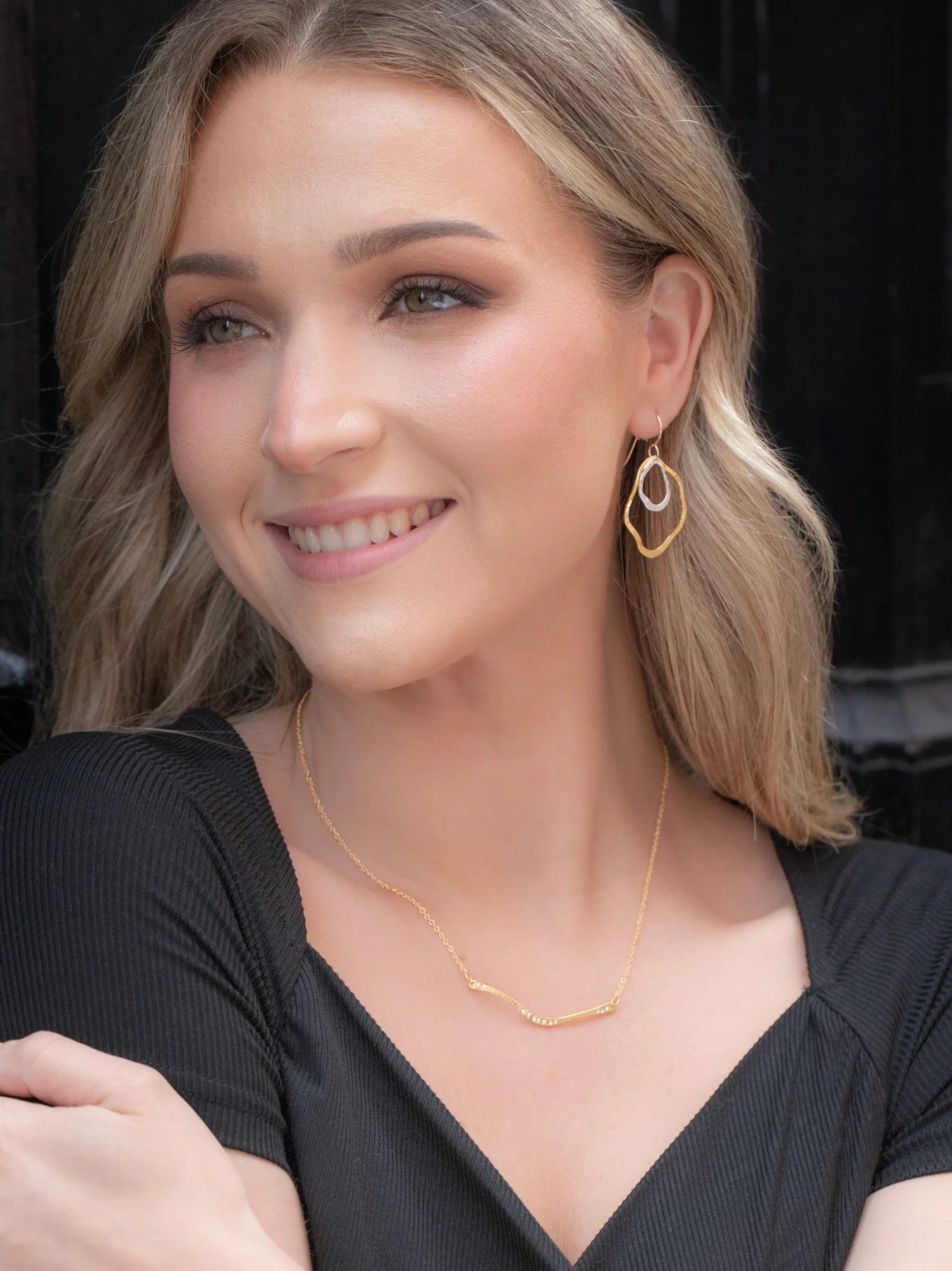 A woman with long blonde hair smiles in a black dress and hypoallergenic gold geometric earrings, wearing the Zara Bar Gold Necklace by Holly Yashi. She stands against a dark background.