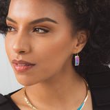 A woman with curly hair wears the Gianna Calypso Collar Necklace by Holly Yashi and matching earrings with iridescent accents. She gazes thoughtfully to the side against a neutral backdrop, her look completed by a gold, hand-hammered pendant.
