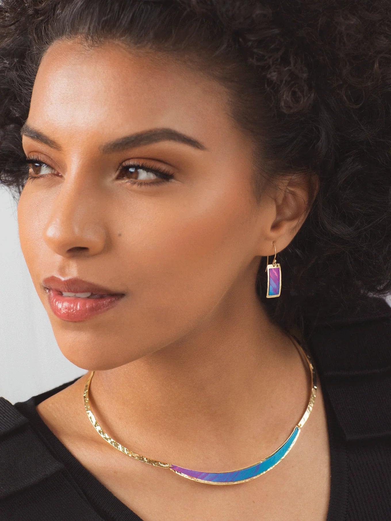 A woman with curly hair wears the Gianna Calypso Collar Necklace by Holly Yashi and matching earrings with iridescent accents. She gazes thoughtfully to the side against a neutral backdrop, her look completed by a gold, hand-hammered pendant.