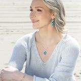 A woman with light hair sits on a beach, smiling and looking to the side. She wears a light blue sweater, Holly Yashi Malia Kai Blue Earrings, a matching necklace, and silver bracelets. Sandy beach and water are in the background.