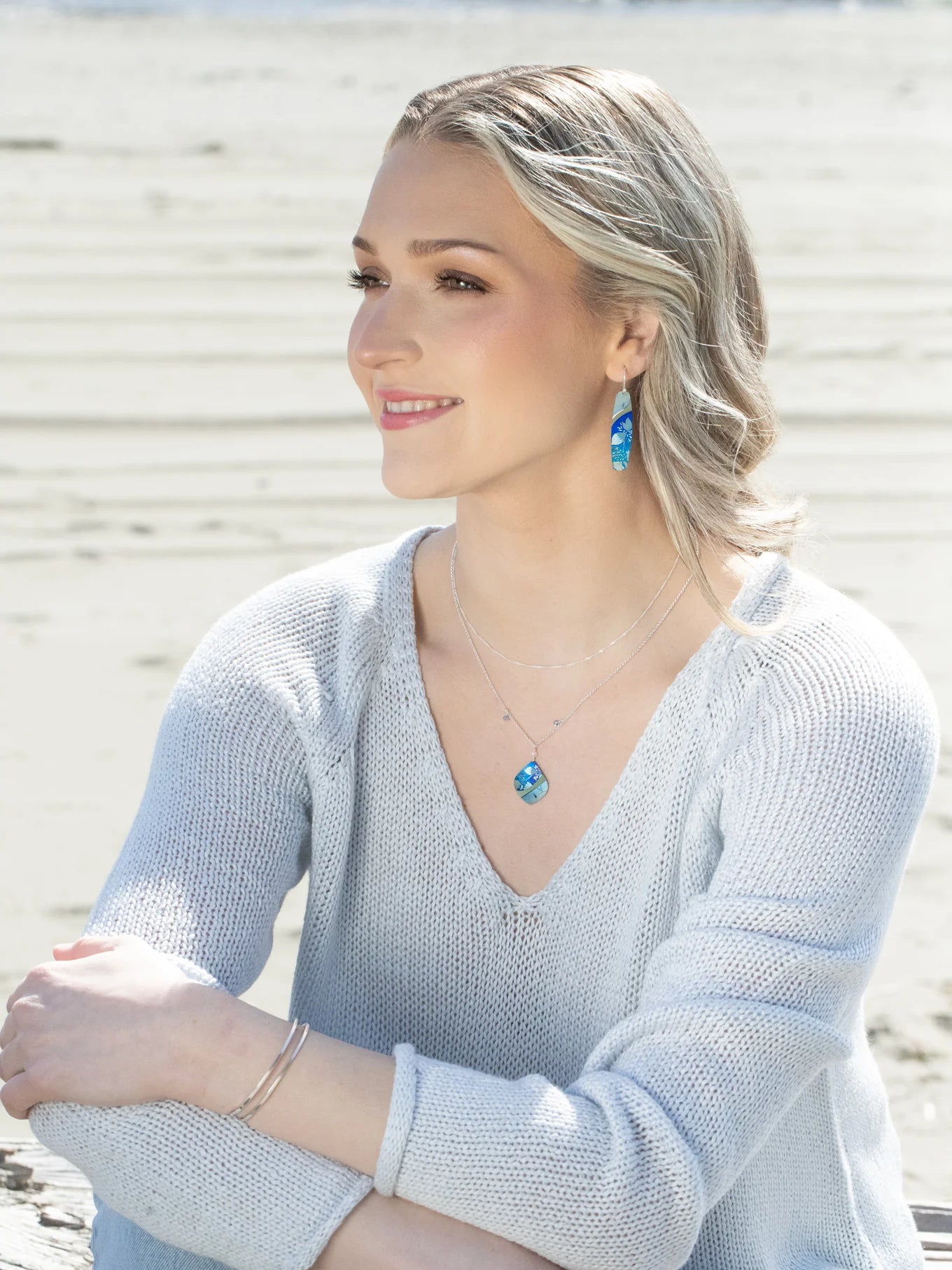 A woman with light hair sits on a beach, smiling and looking to the side. She wears a light blue sweater, Holly Yashi Malia Kai Blue Earrings, a matching necklace, and silver bracelets. Sandy beach and water are in the background.