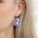 Close-up of a womans ear adorned with Ocean Depth Sea Plum Earrings by Holly Yashi, featuring Austrian crystals in purple and blue. The round silver piece dangles elegantly against her light blonde hair, with a softly blurred background highlighting the earrings beauty.