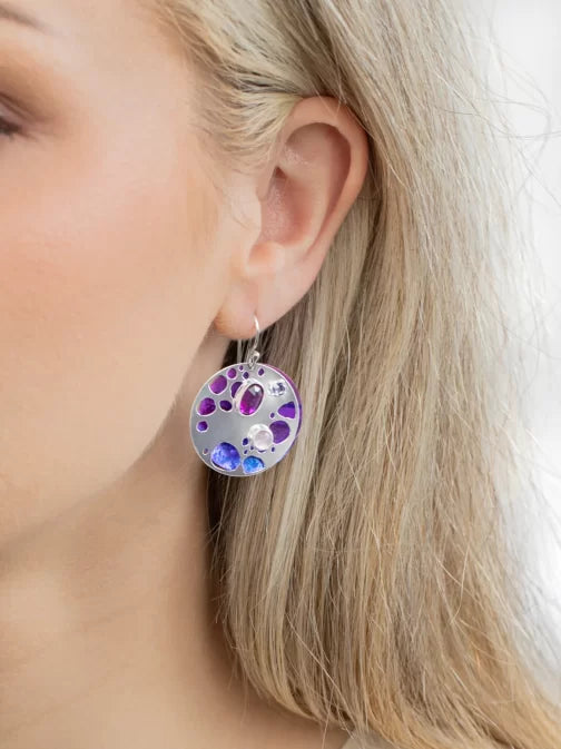 Close-up of a womans ear adorned with Ocean Depth Sea Plum Earrings by Holly Yashi, featuring Austrian crystals in purple and blue. The round silver piece dangles elegantly against her light blonde hair, with a softly blurred background highlighting the earrings beauty.
