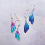 Two pairs of wing-shaped earrings shimmer on a light surface: one is multicolored in pink, teal, and yellow; the other is blue. Both have niobium and 18k gold-filled French wires. These are the vibrant Blue Violet Flutterby Monarch Earrings by Holly Yashi Jewelry.