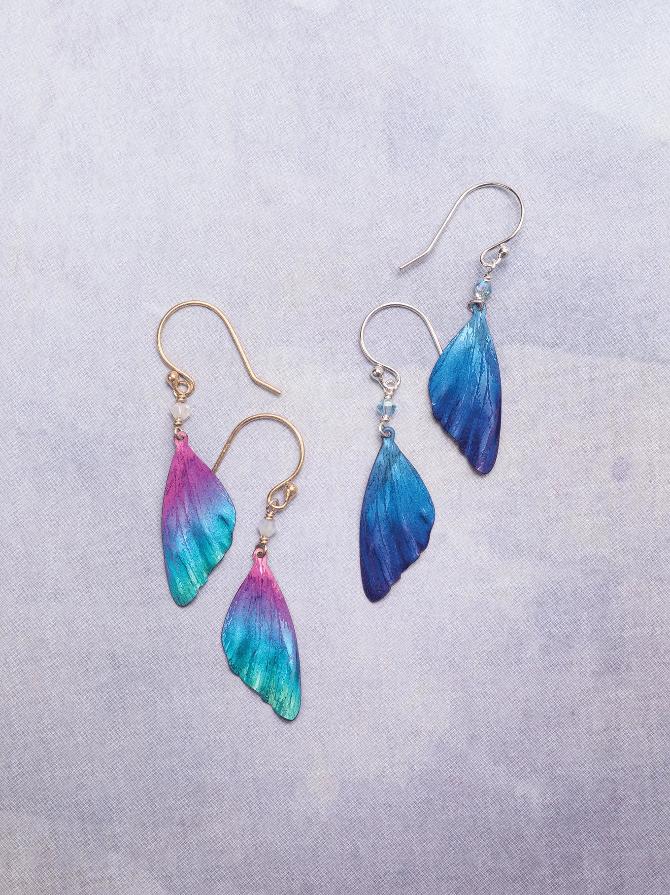 Two pairs of wing-shaped earrings shimmer on a light surface: one is multicolored in pink, teal, and yellow; the other is blue. Both have niobium and 18k gold-filled French wires. These are the vibrant Blue Violet Flutterby Monarch Earrings by Holly Yashi Jewelry.