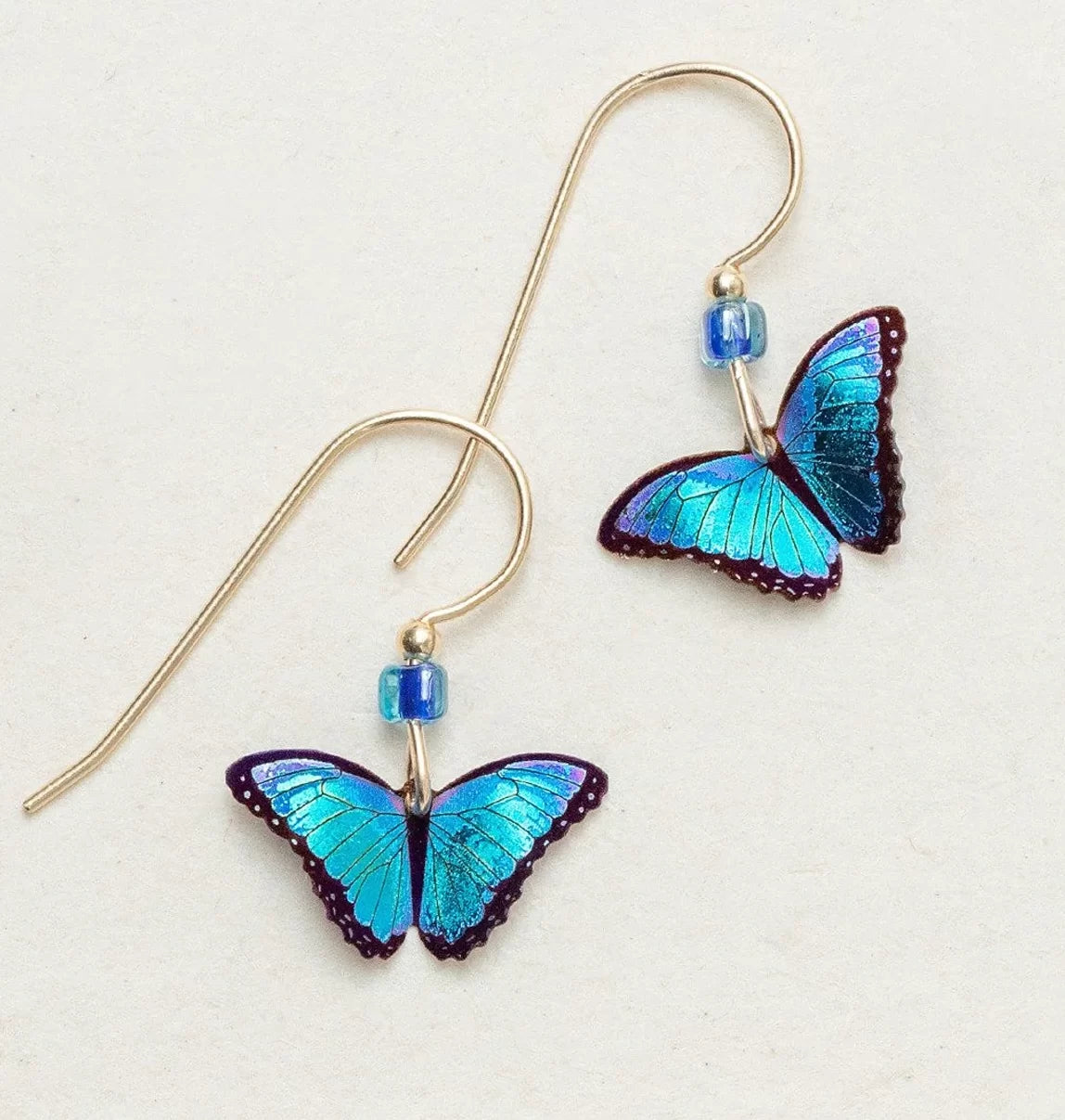 The Blue Petite Bella Butterfly Earrings by Holly Yashi showcase vibrant blue butterfly wings with dark edges, elegantly hanging from thin gold hooks, and feature a small blue bead above each butterfly on an off-white backdrop.