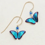 The Blue Petite Bella Butterfly Earrings by Holly Yashi showcase vibrant blue butterfly wings with dark edges, elegantly hanging from thin gold hooks, and feature a small blue bead above each butterfly on an off-white backdrop.