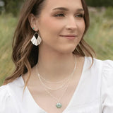 A woman with long brown hair in a white blouse is outdoors, accessorized with hypo-allergenic Silver Ariel Earrings by Holly Yashi and layered necklaces, one showcasing a light blue gemstone. The blurred green backdrop enhances an ocean-inspired tranquility.