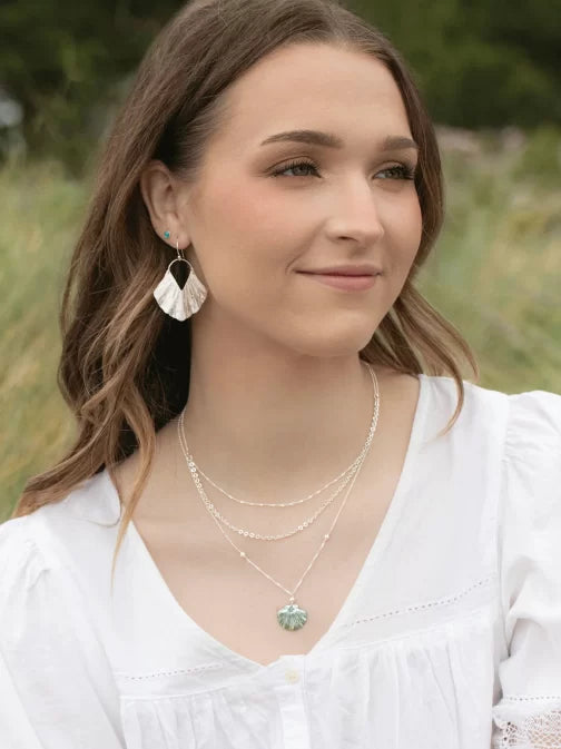 A woman with long brown hair in a white blouse is outdoors, accessorized with hypo-allergenic Silver Ariel Earrings by Holly Yashi and layered necklaces, one showcasing a light blue gemstone. The blurred green backdrop enhances an ocean-inspired tranquility.