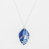 The Blooming Elm Indigo Necklace by Holly Yashi features a silver chain and a blue, leaf-shaped pendant with intricate white botanical designs. This elegant piece of jewelry is shown against a white background.