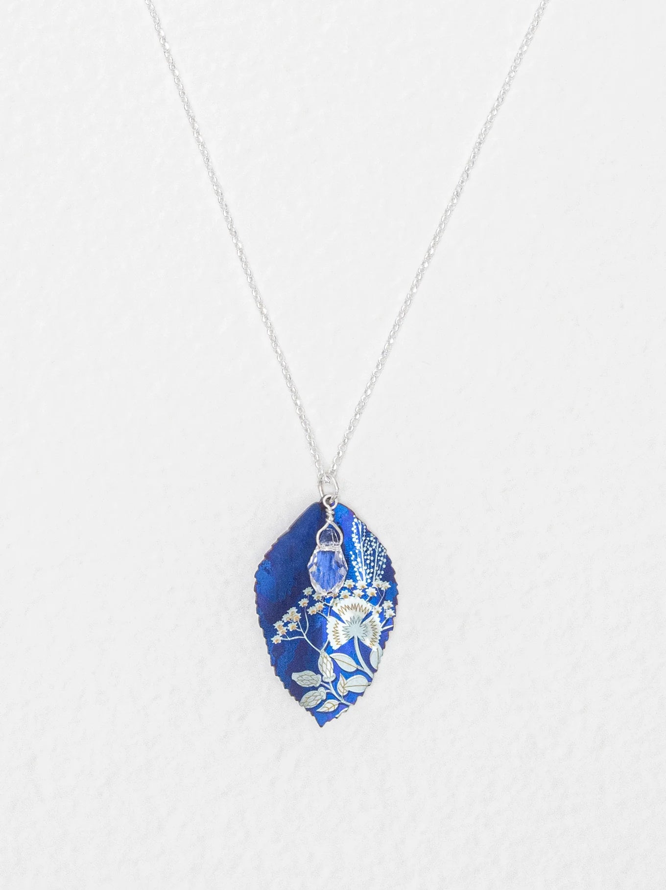 The Blooming Elm Indigo Necklace by Holly Yashi features a silver chain and a blue, leaf-shaped pendant with intricate white botanical designs. This elegant piece of jewelry is shown against a white background.