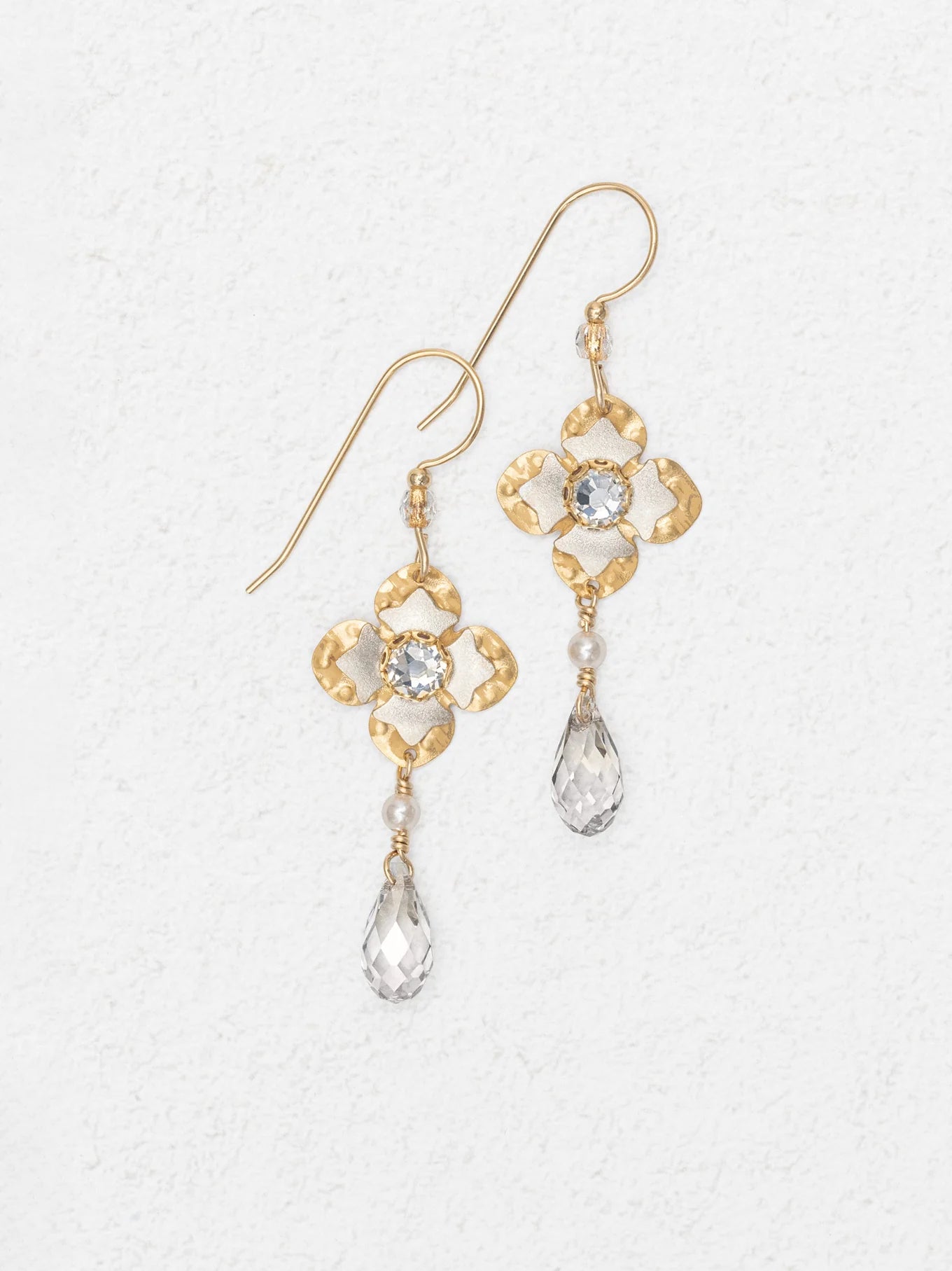 Octavia Gold Silver Earrings by Holly Yashi feature a floral design with a clear central gemstone and teardrop crystal drop, shown on a white textured background.