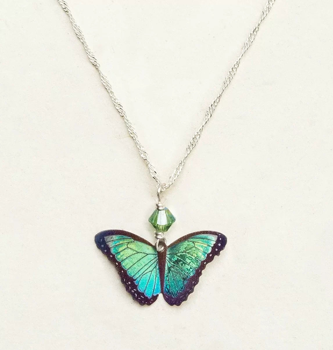 The Green Flash Bella Butterfly Necklace by Holly Yashi features a striking blue and green butterfly pendant on a sterling silver chain, enhanced with a small green crystal bead. Its vibrant wings create an eye-catching centerpiece against the plain background.