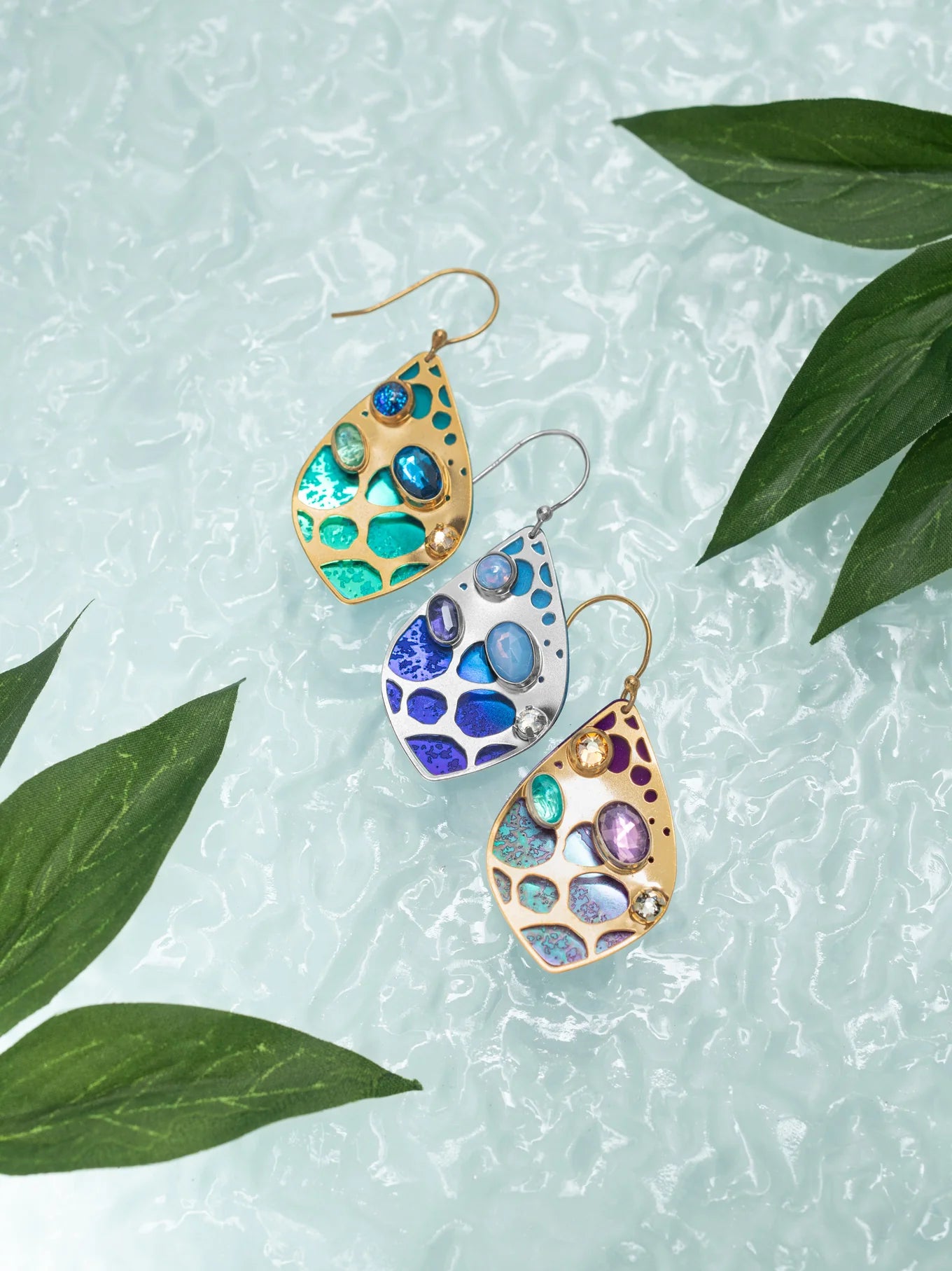 Three Seashore Dreams Purple Earrings by Holly Yashi, featuring bohemian glass accents, rest on a textured light blue surface. Each is encircled by green leaves, adding a natural touch to the composition.