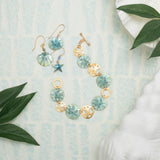 The Sage Oceana Bracelet by Holly Yashi embodies oceanic elegance with niobium, blue shell designs, starfish motifs on a textured surface, and 18k gold-plated details. Green leaves and a white seashell frame the scene, adding charm to this exquisite piece.