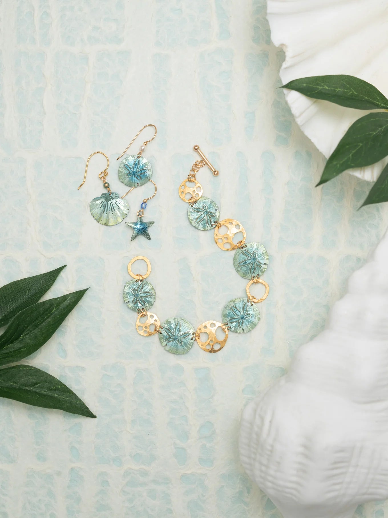 The Sage Oceana Bracelet by Holly Yashi embodies oceanic elegance with niobium, blue shell designs, starfish motifs on a textured surface, and 18k gold-plated details. Green leaves and a white seashell frame the scene, adding charm to this exquisite piece.