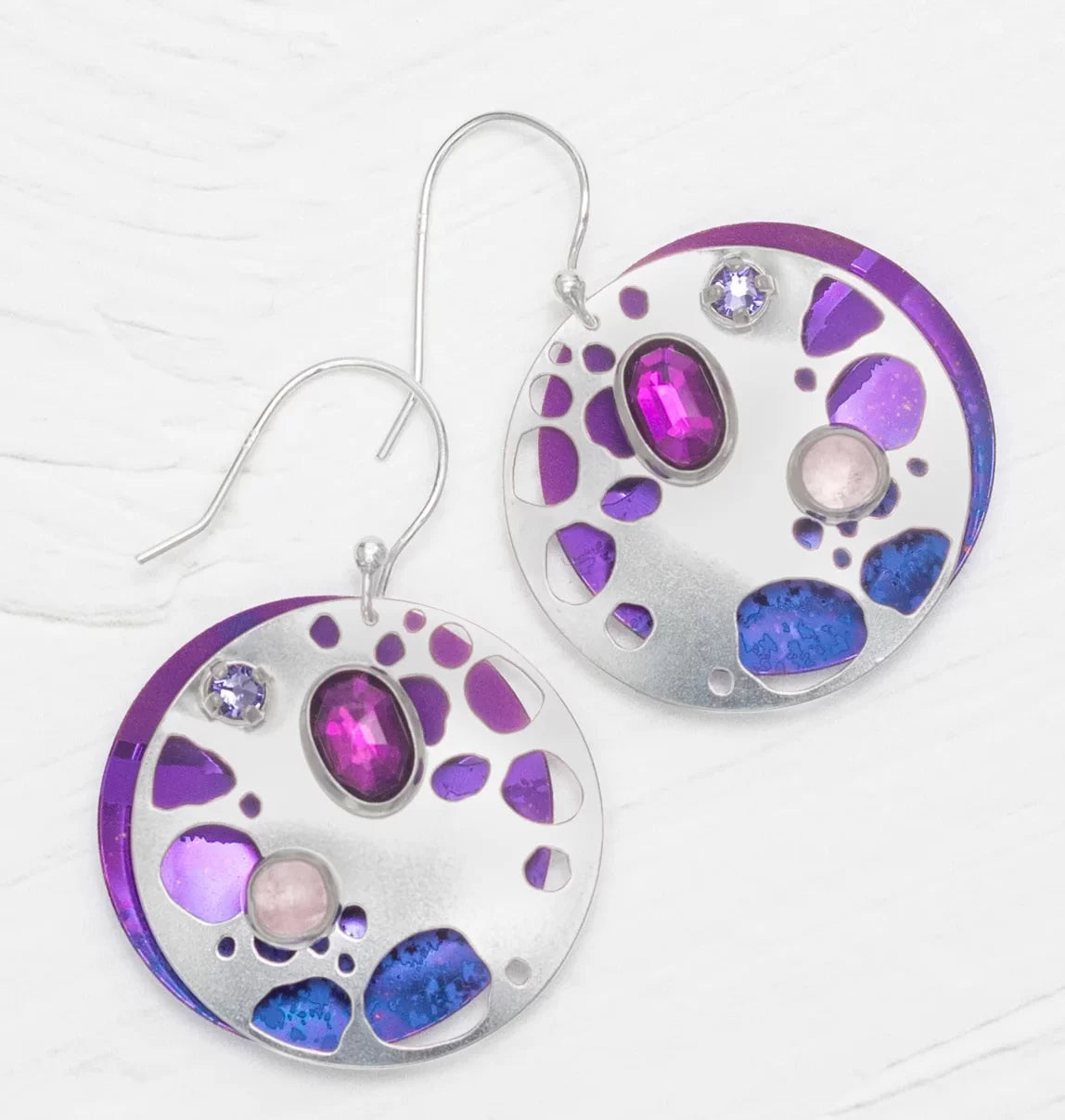 Introducing the Ocean Depth Sea Plum Earrings by Holly Yashi: round silver earrings with abstract cutouts, purple and blue accents, featuring two gemstones—an oval magenta and a round light pink—on textured discs. These hypo-allergenic beauties sparkle with shimmering Austrian crystals.