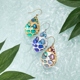 The Seashore Dreams Teal Earrings by Holly Yashi, featuring vibrant Austrian crystals in gold and silver settings, are displayed on a textured light green surface. Lush green leaves surround them, crafting an elegant and natural composition.