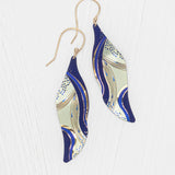 The Turning Leaf Indigo Earrings by Holly Yashi feature wavy, leaf-shaped drops with a blue, gold, and cream abstract design on gold-filled wires, shown on a white textured background.