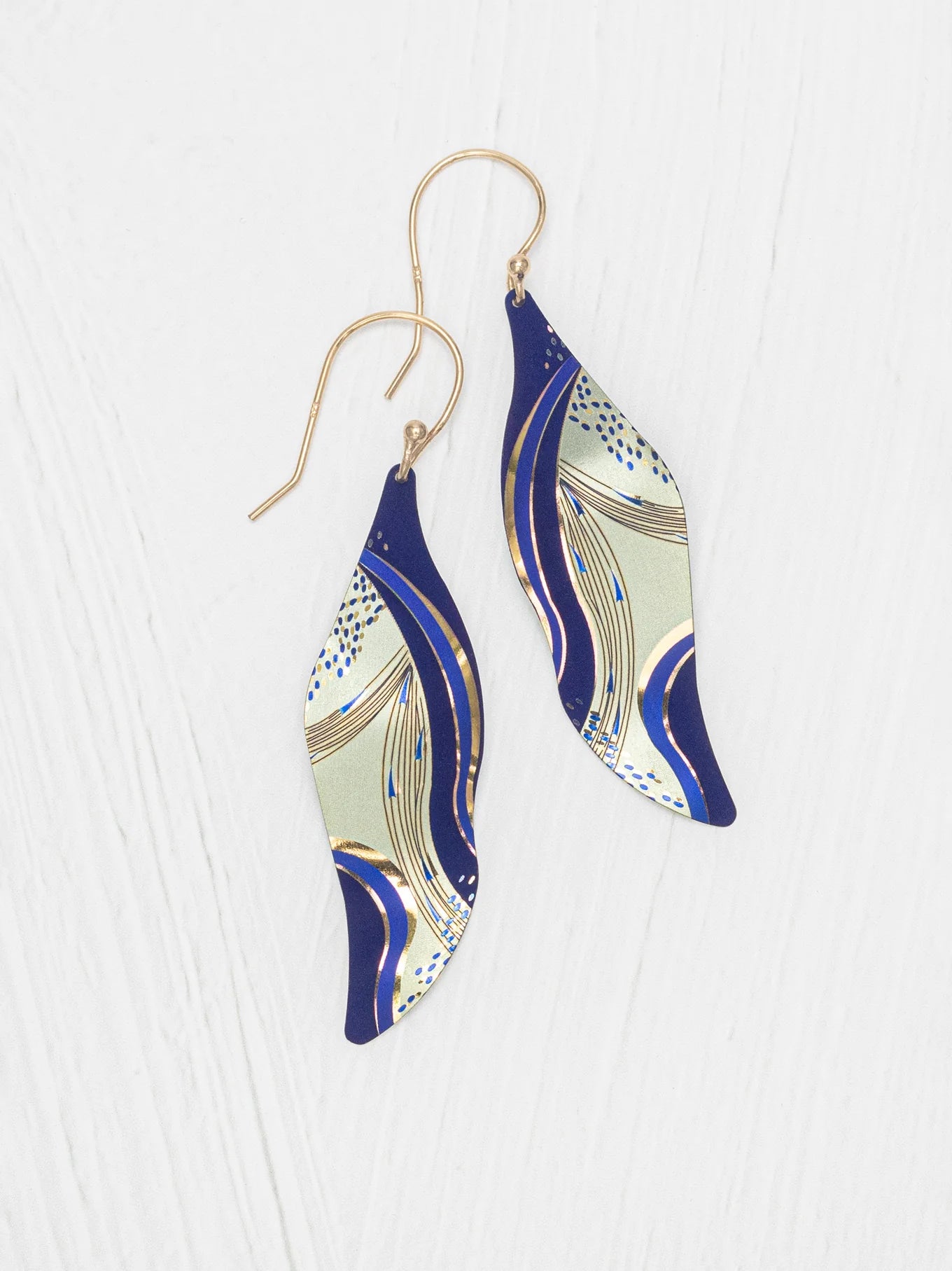 The Turning Leaf Indigo Earrings by Holly Yashi feature wavy, leaf-shaped drops with a blue, gold, and cream abstract design on gold-filled wires, shown on a white textured background.