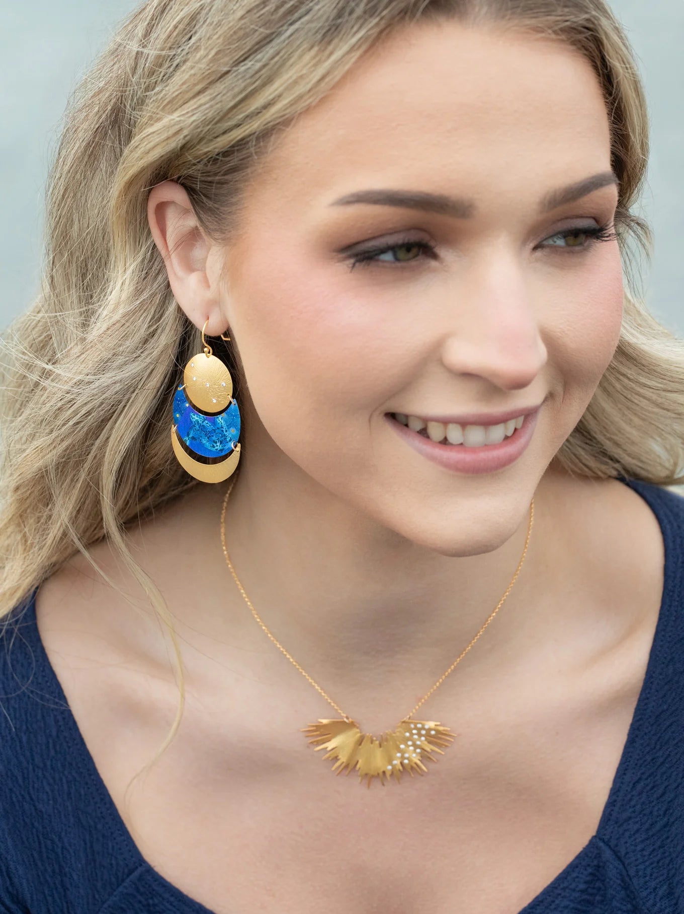 A woman with wavy blonde hair smiles, wearing a navy top and the Holly Yashi Aster Necklace-Gold Necklace, which features a sunburst design, along with large gold dangle earrings accented with shimmering blue crystal stones.
